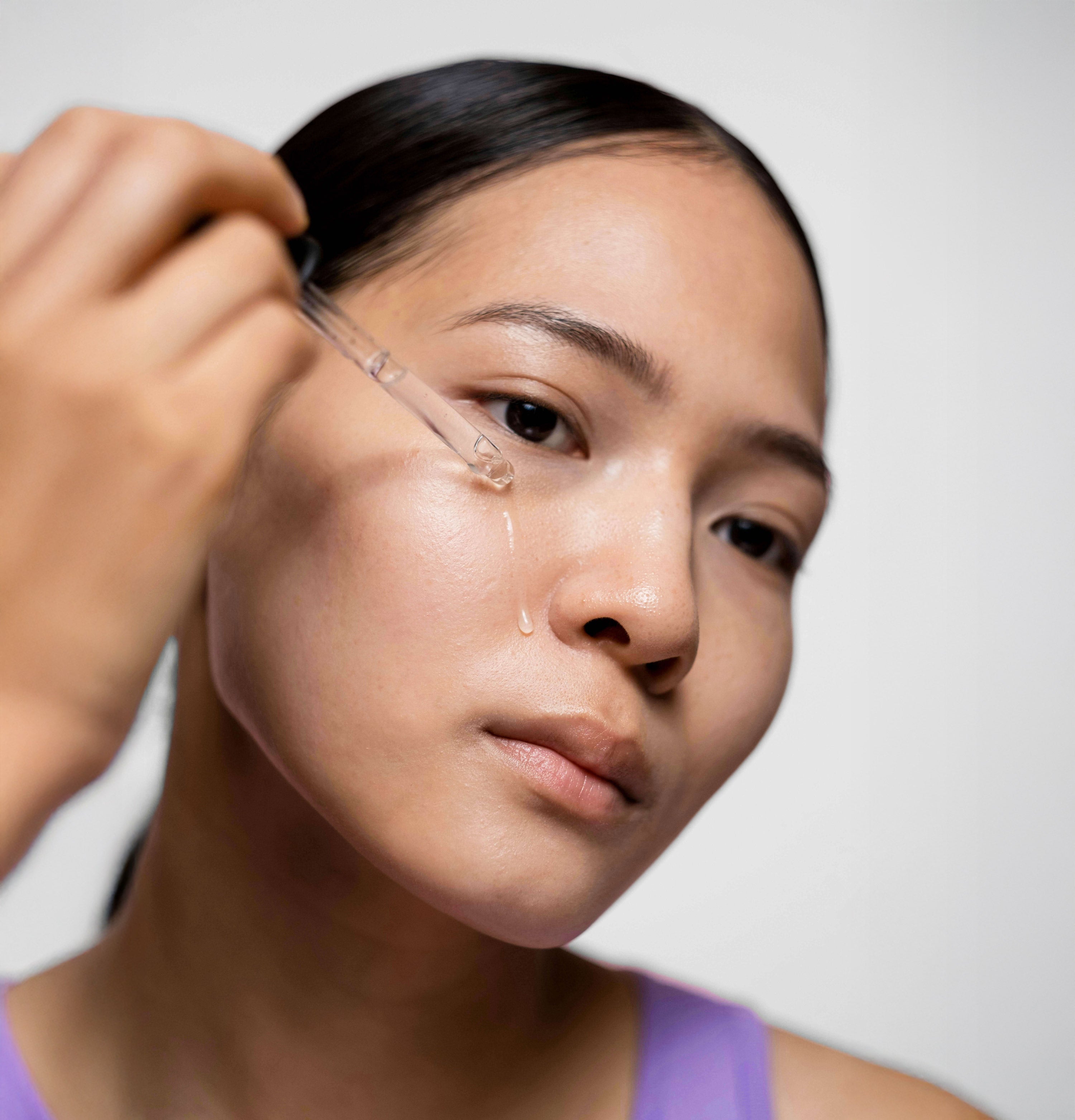 Person applying a dropper of liquid to their face against a neutral background